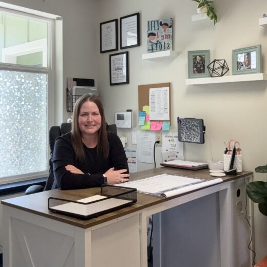 A woman sits at a desk in a neatly organized office with framed photos and documents on the wall, shelves with decor, and office supplies on the desk.