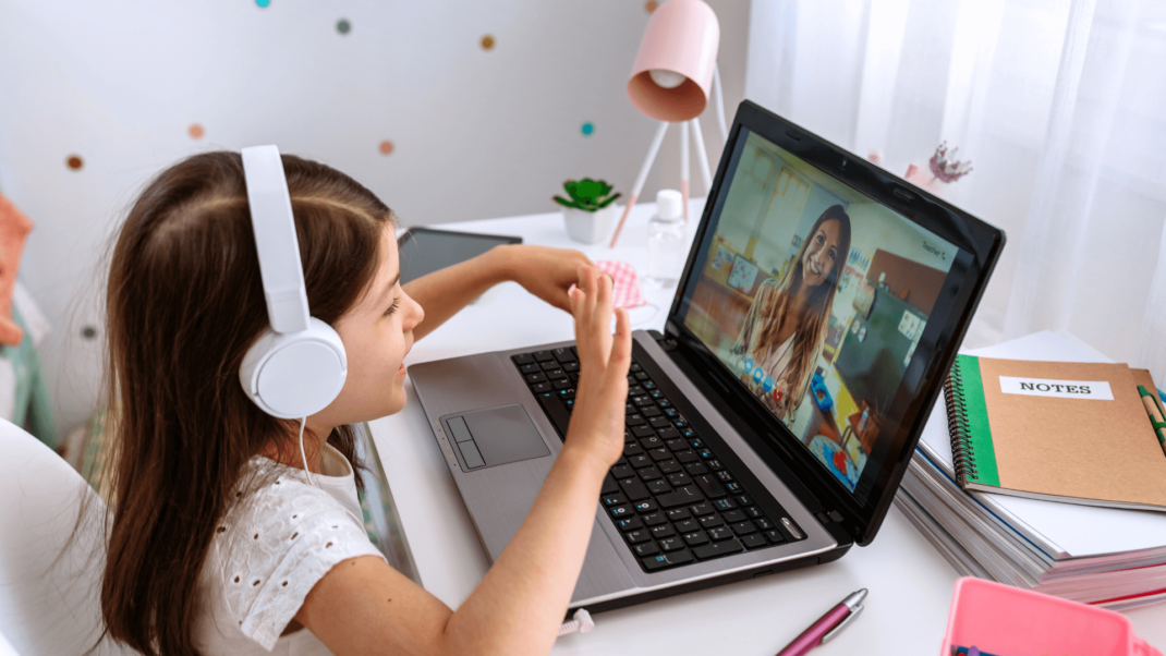 A young girl wearing headphones sits at a desk, waving at a woman on a video call displayed on her laptop screen.
