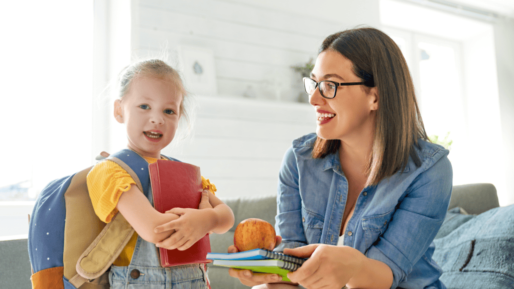 A young girl with a backpack holds a book while an adult woman hands her notebooks and an apple inside a brightly lit room.
