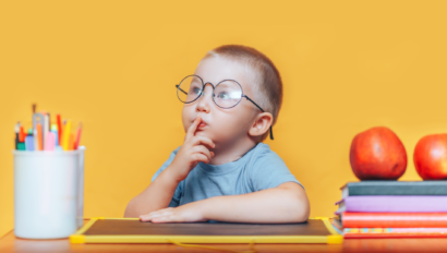 A young child wearing round glasses sits at a desk, holding a finger to their lips, surrounded by books, apples, and colored pencils, against an orange background.