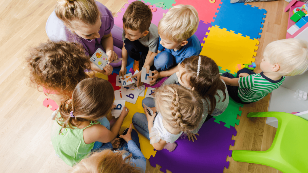 A group of young children sit in a circle on colorful foam mats, learning with letter cards, while an adult supervises.