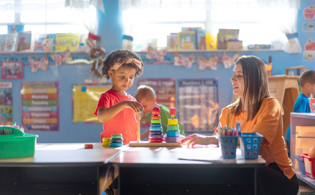 A young child stacks colorful rings on a peg while a teacher watches attentively in a brightly lit classroom.