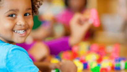 A young child smiling at the camera while sitting at a table with colorful building blocks; other children play in the background.