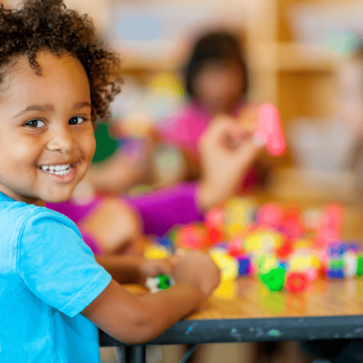 A young child smiling at the camera while sitting at a table with colorful building blocks; other children play in the background.