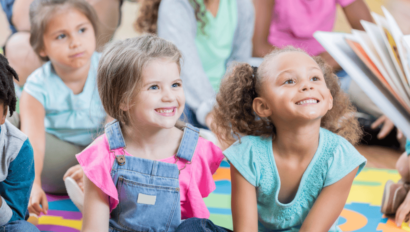 A group of young children sit on a colorful mat, smiling and looking toward an open book being read to them.