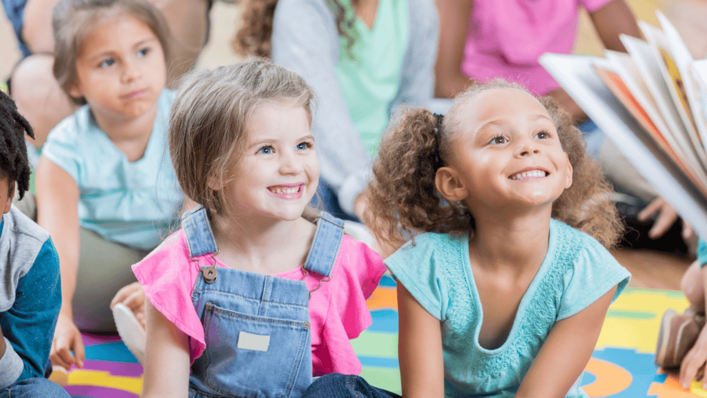 A group of young children sit on a colorful mat, smiling and looking toward an open book being read to them.