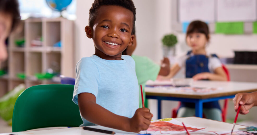 Smiling young boy in a classroom, painting with a brush at a table, with other children working in the background.